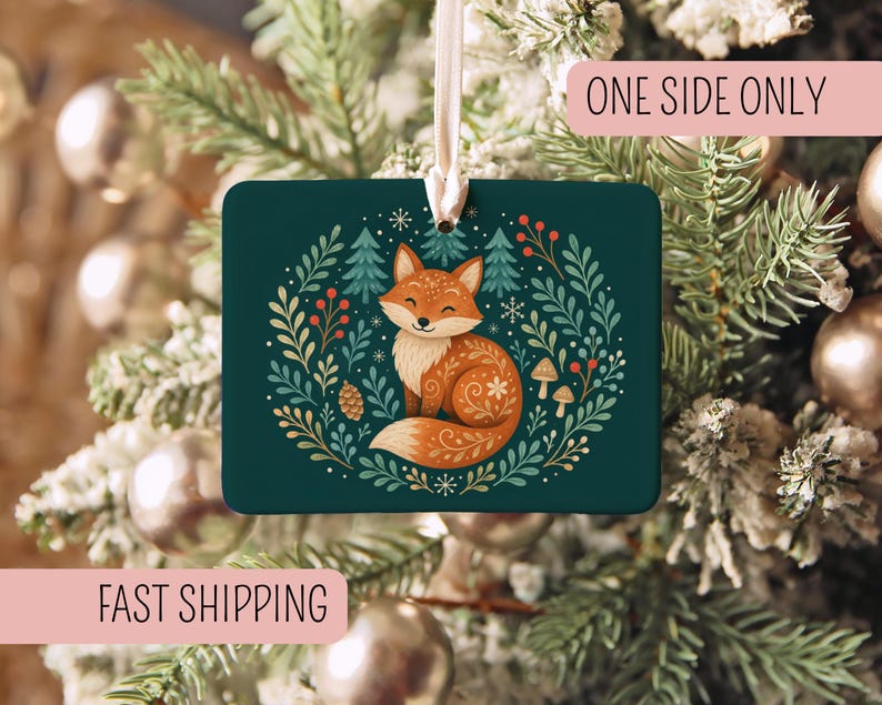 May include: A rectangular dark green ornament featuring a central illustration of a fox surrounded by a wreath of pine trees, berries, mushrooms, and snowflakes. The ornament hangs from a white ribbon and includes the text ONE SIDE ONLY.