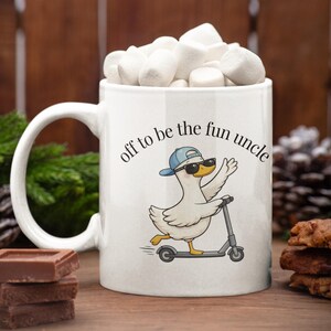 May include: White ceramic mug filled with marshmallows, featuring a cartoon duck riding a scooter. The duck wears sunglasses and a blue cap, with the text "off to be the fun uncle" in a circular arc above the duck.