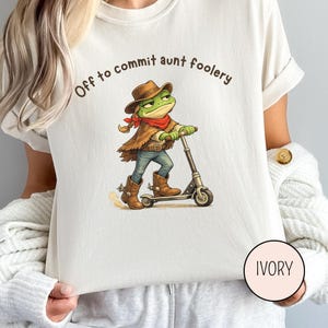 May include: Ivory-colored t-shirt featuring a cartoon frog in a cowboy hat and boots riding a scooter. The frog wears a red bandana and brown vest. Text above the frog reads "Off to commit aunt foolery."