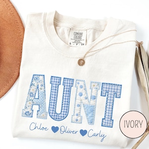 May include: Ivory-colored t-shirt with the word "AUNT" in blue patchwork letters. The letters feature floral, polka dot, and gingham patterns. Below the word are names and blue heart symbols. A gold necklace is draped across the shirt.