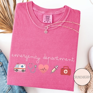 以下が含まれることがあります： ピンク色のComfort ColorsのTシャツ。筆記体で「emergency department」と書かれています。救急車、聴診器、心拍、注射器、救急箱などの医療をテーマにしたグラフィックが特徴です。花のペンダントが付いたシルバーのネックレスがシャツの上にかけられています。