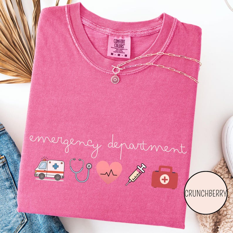 以下が含まれることがあります： ピンク色のComfort ColorsのTシャツ。筆記体で「emergency department」と書かれています。救急車、聴診器、心拍、注射器、救急箱などの医療をテーマにしたグラフィックが特徴です。花のペンダントが付いたシルバーのネックレスがシャツの上にかけられています。