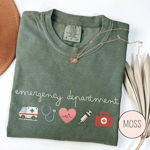以下が含まれることがあります： モスグリーンのTシャツ。白の筆記体で「emergency department」と書かれています。救急車、聴診器、心臓、注射器、救急箱など、医療をテーマにしたグラフィックが特徴です。金のネックレスがTシャツの上にあります。