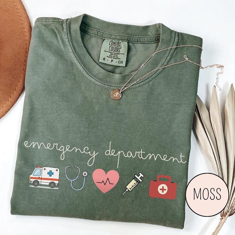以下が含まれることがあります： モスグリーンのTシャツ。白の筆記体で「emergency department」と書かれています。救急車、聴診器、心臓、注射器、救急箱など、医療をテーマにしたグラフィックが特徴です。金のネックレスがTシャツの上にあります。