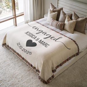 May include: Cream-colored throw blanket with the word "engaged" in a script font, along with the names "JESSICA & MARC" and the date "12.10.2025". A black heart graphic is also featured. The blanket has a fringed edge.