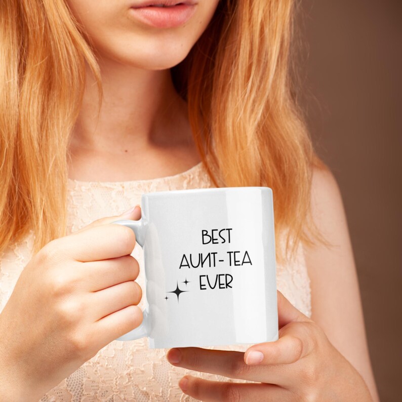 May include: White ceramic mug with the text "BEST AUNT-TEA EVER" in black. The mug has a standard handle and a small star graphic. The mug is held by a person with long, light-colored hair.