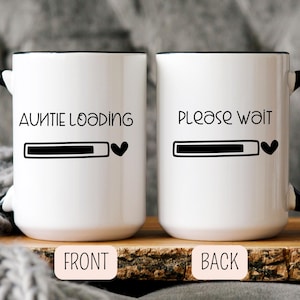 May include: Two white ceramic mugs with black rims and handles. One mug reads "AUNTIE LOADING" with a loading bar and heart symbol. The other mug says "PLEASE WAIT" with a loading bar and heart. The mugs are on a wooden surface.
