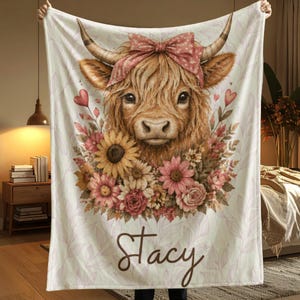 May include: A cream-colored blanket featuring a Highland cow illustration. The cow has a pink bow, surrounded by flowers and hearts. The name "Stacy" is written in a cursive font at the bottom. The blanket has a floral pattern.