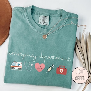 以下が含まれることがあります： 「emergency department」の文字が白で書かれた、薄い緑色のTシャツ。救急車、聴診器、心臓、注射器、救急箱のイラストが描かれています。ペンダント付きのゴールドのネックレスがシャツの上にかけられています。