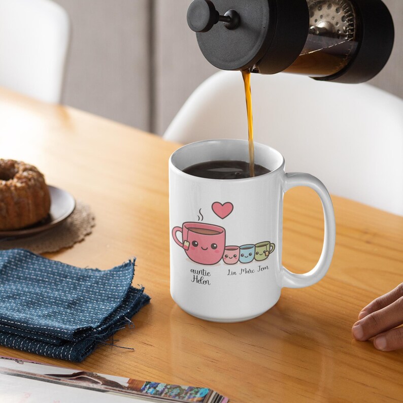 May include: A white ceramic mug with a cartoon design of a pink teacup, a heart, and three smaller cups. The mug has the text "auntie Helen" and "Lin Mae Jem". Coffee is being poured into the mug from a French press.