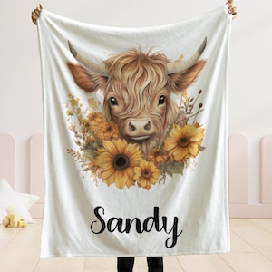 May include: White blanket featuring a watercolor illustration of a Highland cow with sunflowers. The cow has brown fur and large horns. The name "Sandy" is written in black script at the bottom. The blanket is held up by a person.