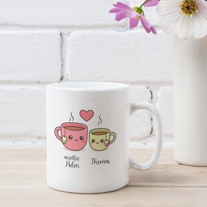 May include: White ceramic mug featuring a pink and green cartoon teacup design with steam and a heart. The mug has the text "auntie Helen" and "Thomas" below the teacups. A perfect gift for tea lovers.