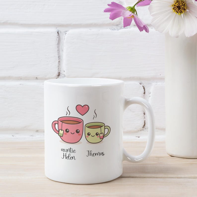 May include: White ceramic mug featuring a pink and green cartoon teacup design with steam and a heart. The mug has the text "auntie Helen" and "Thomas" below the teacups. A perfect gift for tea lovers.