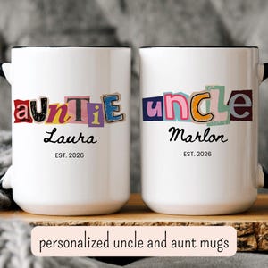 May include: Two white ceramic mugs with black rims and handles. One mug says "auntie Laura EST. 2026" in a colorful, cut-out style. The other says "uncle Marlon EST. 2026". The mugs are on a wooden surface.