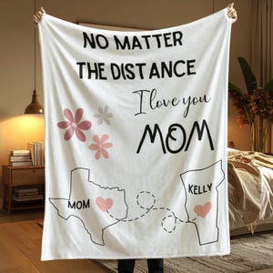 May include: White fleece blanket with the text "NO MATTER THE DISTANCE I love you MOM" in black. Features pink floral accents, state outlines with hearts, and the names "MOM" and "KELLY".