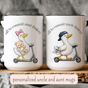 May include: Two white ceramic mugs with black rims. One mug features a cartoon duck with a pink bow, holding a teddy bear, and riding a scooter. The other mug shows a duck wearing a cap, holding sneakers, and riding a scooter. Text reads "off to commit aunt foolery" and "off to commit uncle foolery".