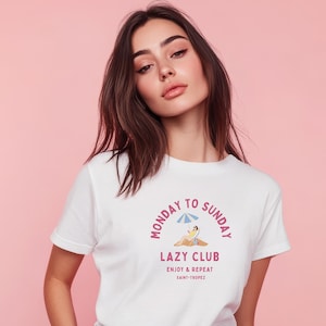 May include: White t-shirt with a graphic that reads "Monday to Sunday Lazy Club Enjoy & Repeat Saint-Tropez". The graphic features a person relaxing under an umbrella. The model is wearing blue jeans.