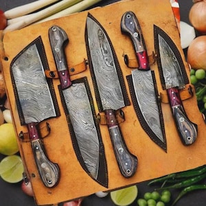 May include: A set of six Damascus steel kitchen knives with wooden handles and red accents, displayed in a brown leather sheath. The blades have a unique, wavy pattern. The knives are arranged on a wooden surface.