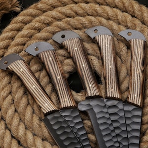May include: Five handcrafted knives with textured black blades and wooden handles. The handles feature a light and dark wood grain pattern with small brass rivets. The knives are arranged on a coiled rope background.