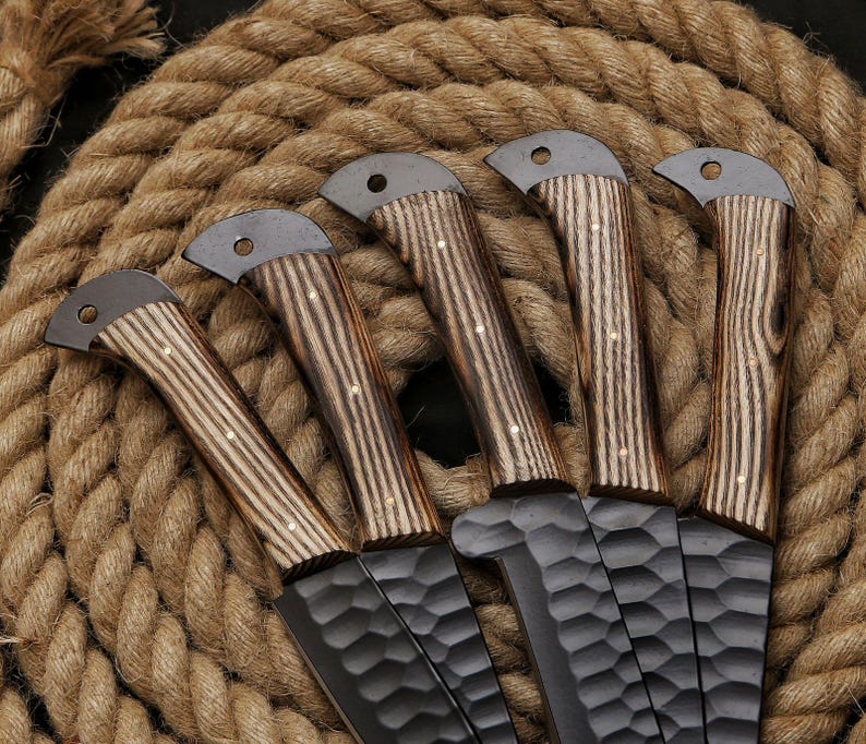 May include: Five handcrafted knives with textured black blades and wooden handles. The handles feature a light and dark wood grain pattern with small brass rivets. The knives are arranged on a coiled rope background.