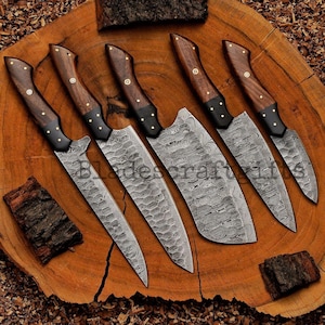 May include: Six handcrafted knives with wooden handles and patterned blades are arranged on a wooden surface. The blades have a unique textured design. The handles are dark wood with brass rivets and black bolsters.
