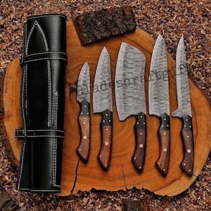 May include: A set of six Damascus steel knives with wooden handles and a black leather carrying case. The blades have a distinctive patterned design. The knives are arranged on a wooden surface.