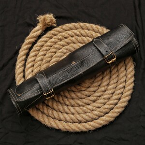 May include: A rolled black leather knife roll with white stitching and brass buckles, resting on a coiled rope. The leather has a textured appearance. The rope is a light brown color, and the background is black.