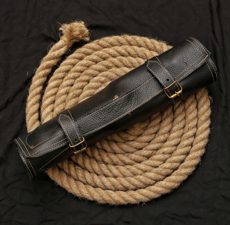 May include: A rolled black leather knife roll with white stitching and brass buckles, resting on a coiled rope. The leather has a textured appearance. The rope is a light brown color, and the background is black.