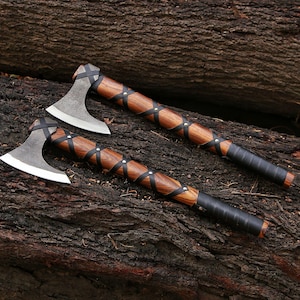 May include: Two Viking-style axes with wooden handles and metal blades. The handles are wrapped with black leather straps and silver rivets. The axes are resting on a dark brown, textured surface, possibly a log.