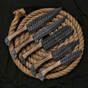 May include: A set of six black kitchen knives with textured blades and wooden handles. The knives are arranged on a coiled rope, showcasing their craftsmanship and design. The blades have a unique hammered texture.