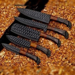 May include: An assortment of five black chef knives with textured blades and wooden handles. The handles have a dark finish with a lighter wood accent near the blade. The knives are arranged on a bed of wood shavings.