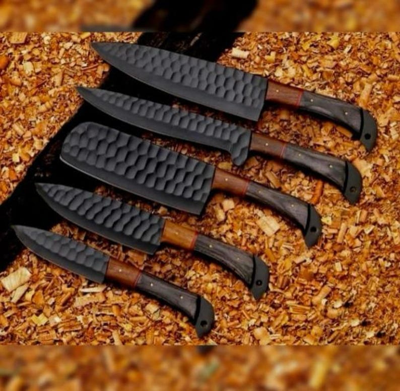May include: An assortment of five black chef knives with textured blades and wooden handles. The handles have a dark finish with a lighter wood accent near the blade. The knives are arranged on a bed of wood shavings.