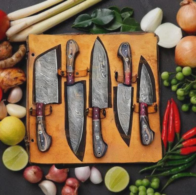 May include: A set of five stainless steel knives with wooden handles and leather sheaths, displayed on a tan leather surface. The blades have a unique patterned design. Various vegetables and herbs surround the knives, including onions, limes, and chili peppers.