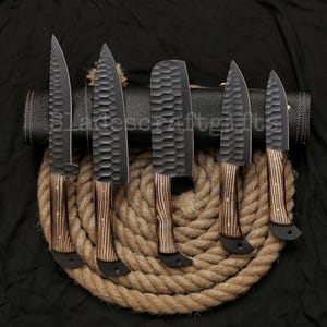May include: A set of six black kitchen knives with textured blades and wooden handles. The knives are arranged on a coil of rope, with a black leather knife roll in the background. The blades have a hammered finish.