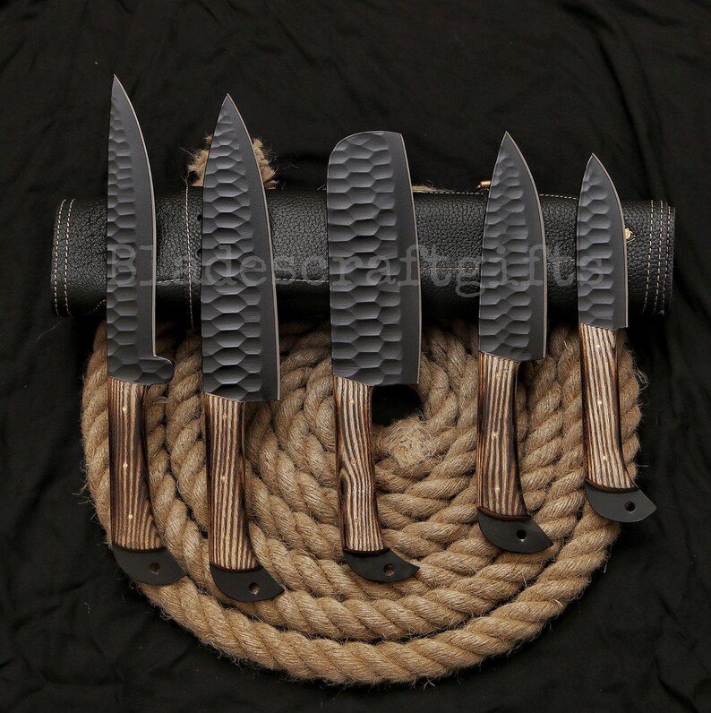 May include: A set of six black kitchen knives with textured blades and wooden handles. The knives are arranged on a coil of rope, with a black leather knife roll in the background. The blades have a hammered finish.