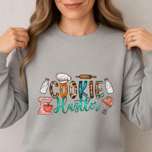 May include: A light gray sweatshirt with the words "COOKIE Hustler" in colorful, whimsical lettering. The design includes illustrations of a chef's hat, rolling pin, milk bottles, and a mixer, perfect for a baker.