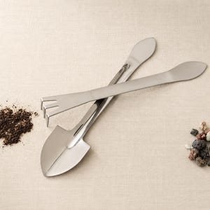 May include: Two silver-toned metal gardening tools, one shaped like a small shovel and the other like a miniature rake, are crossed. A small pile of dark brown soil is positioned to the left, and a cluster of small, multi-coloured stones is to the right.