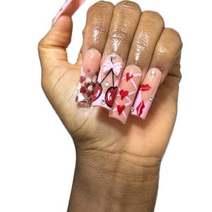 May include: A hand displaying long, pink acrylic nails with various designs. One nail features red and white hearts, while others showcase cherries, bows, hearts, and lips. Each nail is adorned with small, sparkling embellishments.