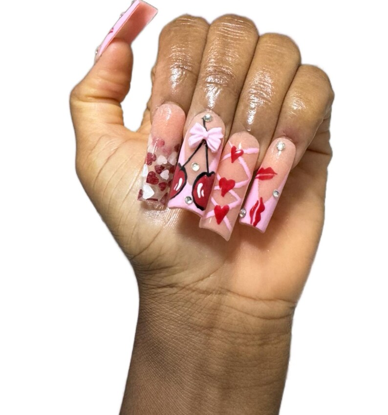 May include: A hand displaying long, pink acrylic nails with various designs. One nail features red and white hearts, while others showcase cherries, bows, hearts, and lips. Each nail is adorned with small, sparkling embellishments.