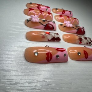 May include: A collection of artificial nails with various designs. Some nails feature a cherry design with pink and red accents, while others have a lip design with rhinestones. The nails are in shades of pink, peach, and red.
