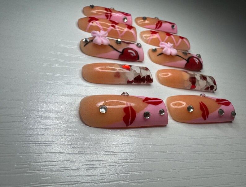 May include: A collection of artificial nails with various designs. Some nails feature a cherry design with pink and red accents, while others have a lip design with rhinestones. The nails are in shades of pink, peach, and red.