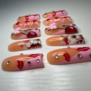 May include: A collection of ten artificial nails, each with a unique design. The nails feature a combination of pink, peach, and red colors, with accents like rhinestones, hearts, and floral details. The nails are designed for cosmetic use.