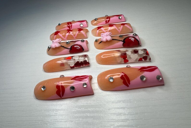 May include: A collection of ten artificial nails, each with a unique design. The nails feature a combination of pink, peach, and red colors, with accents like rhinestones, hearts, and floral details. The nails are designed for cosmetic use.