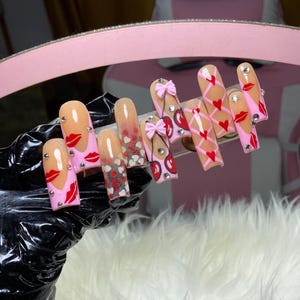 May include: A set of long, square-shaped artificial nails. The nails feature various designs, including red lips, hearts, bows, and cherry designs. Some nails have a gradient effect, transitioning from a nude color to a pink hue. Rhinestones are also incorporated into the designs.