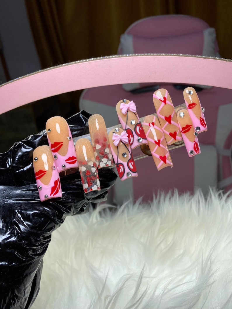 May include: A set of long, square-shaped artificial nails. The nails feature various designs, including red lips, hearts, bows, and cherry designs. Some nails have a gradient effect, transitioning from a nude color to a pink hue. Rhinestones are also incorporated into the designs.