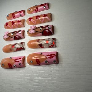 May include: A collection of ten artificial nails with various designs. Some nails feature pink and peach tones with red lip and bow accents, while others have glitter and rhinestone embellishments. The nails are arranged on a light gray surface.