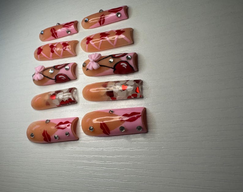 May include: A collection of ten artificial nails with various designs. Some nails feature pink and peach tones with red lip and bow accents, while others have glitter and rhinestone embellishments. The nails are arranged on a light gray surface.