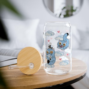 May include: A clear glass can-shaped tumbler with a bamboo lid and straw. The tumbler features cartoon blue cats, clouds, and hearts. The lid and straw are made of natural bamboo. The tumbler is on a wooden table.