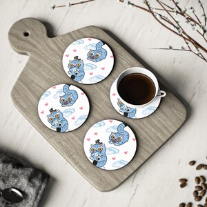 May include: Four round coasters with a playful cat illustration. The coasters display a light blue cat with yellow eyes, clouds, and pink hearts on a white backdrop. A cup of coffee rests on one of the coasters, adding to the scene.