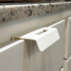 May include: A white cabinet pull handle with a rectangular design and a cutout for gripping. The handle is mounted on a white cabinet door, with a granite countertop and wooden cutting board in the background.